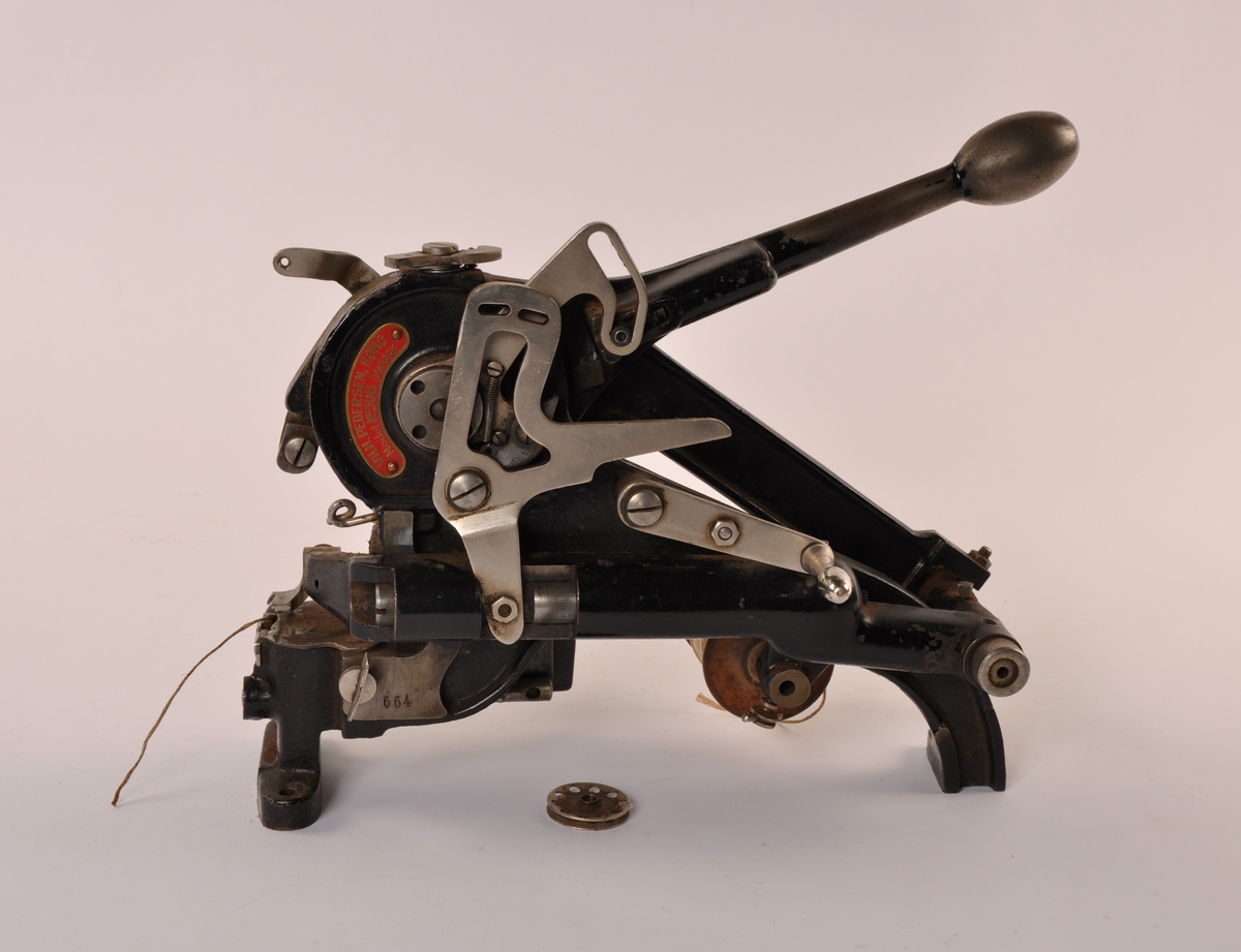 Hånddrevet symaskin i metall drevet av en spak. Til å montere fast på underlag. Tresnelle med snøre.  Løs metallboldt med fjær.