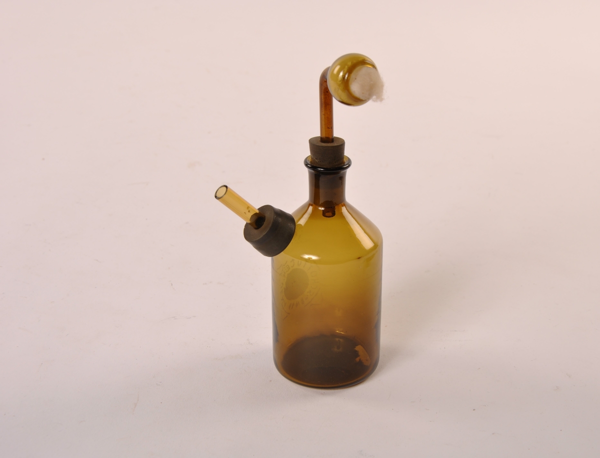 Flaske med avsmalet tut og en avsmalet utstiikkende åpen sylinder. Løs bøyd sylinder festet med gummipropp. Flasken er i brunt glass.