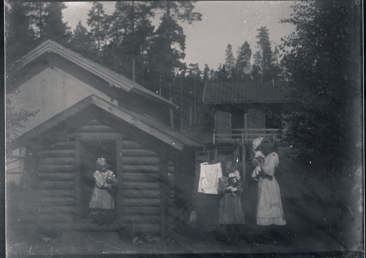 Tre jenter som alle holder på dukker. Jentene er antatt å være tre av døtrene til bestyrer Rydgren på Kistefos. Minste jenta står i døråpningen til et lite, laftet hus. I bakgrunnen er et uthus eller skjul med to åpne dører.