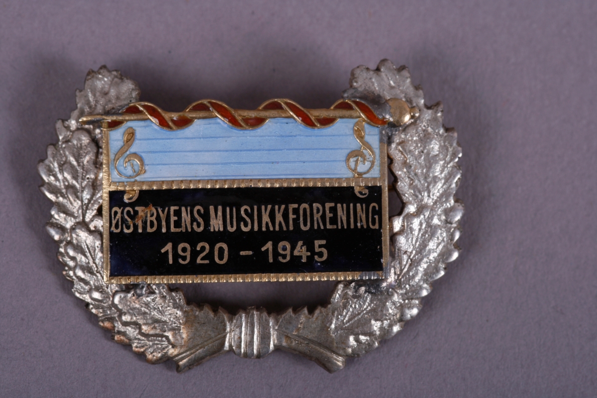 Spenne med sløyfe. Påskrevet Østbyens Musikkforening. 1920-1945.
Fane med takstav, g-nøkler til pynt. Omgitt av laurbærkrans.