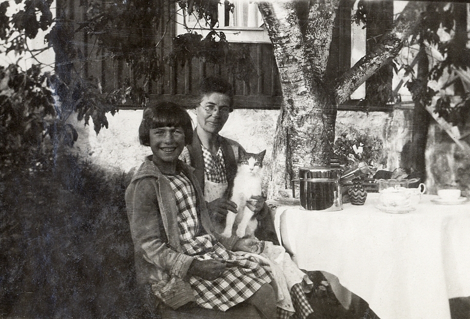 En flicka och en kvinna med en katt i famnen sitter vid ett kaffebord i en trädgård.
Under fotot text: "Hällevik, 1929".