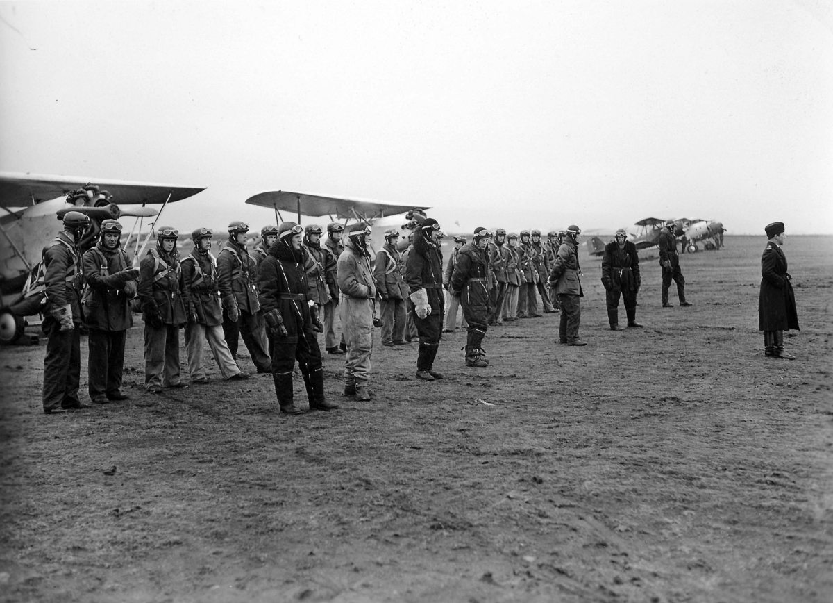 Militära flygare samlade på ett flygfält för ordergivning under 1930-talet. Omkring 25 män framför flygplan S 6. Längst till höger står Nils Söderberg. 1930-tal.