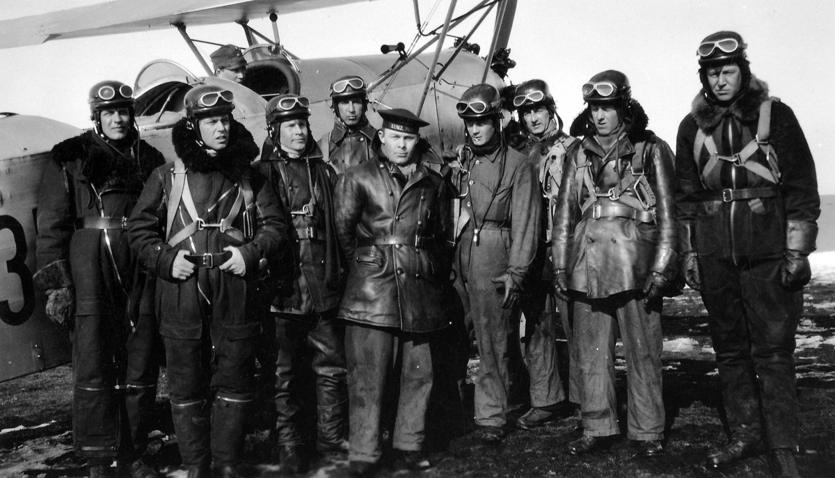 Grupporträtt av nio militärer framför ett flygplan. Åtta män i flygardräkt och en man ur marinen. En ytterligare man sitter i flygplanet. Vintertid, 1930-tal.