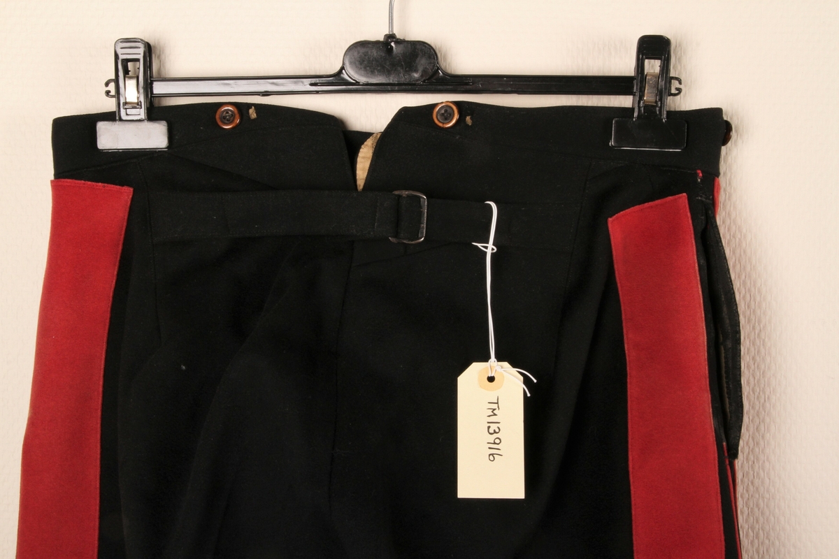 Uniformsbukse i svart ullstoff, med to brede, røde renner i hver side. Spensel i midjen midt bak. 