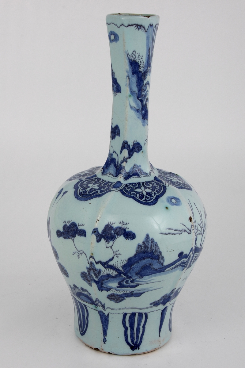 Heksagonal klubbeformet vase med kinesiskinspirert motiv med figur i landskap på både korpus og vasehals. En europeisk versjon av lingzhi/ ruyi-ornamentikk rundt vasehalsen på toppen av korpus.  Palmeblader nederst på vasen under hovedmotivet.