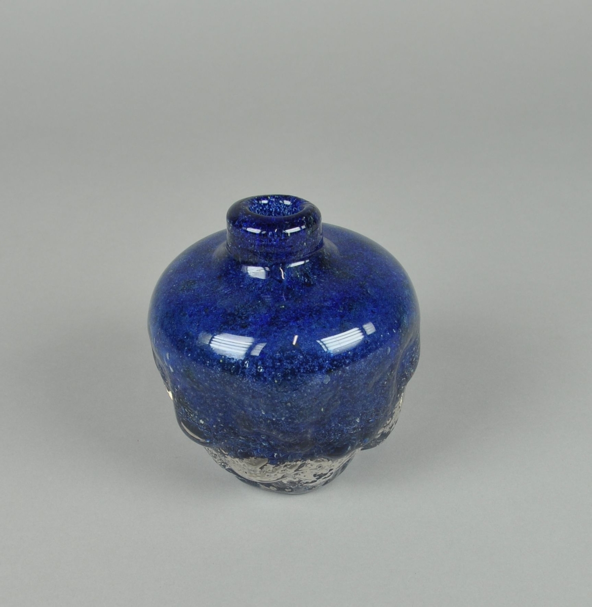 Munnblåst vase av blått glass med klart overfangsglass. Rund form med lav hals. Vasen er farget blå og har boblende tekstur på nedre del av korpus.