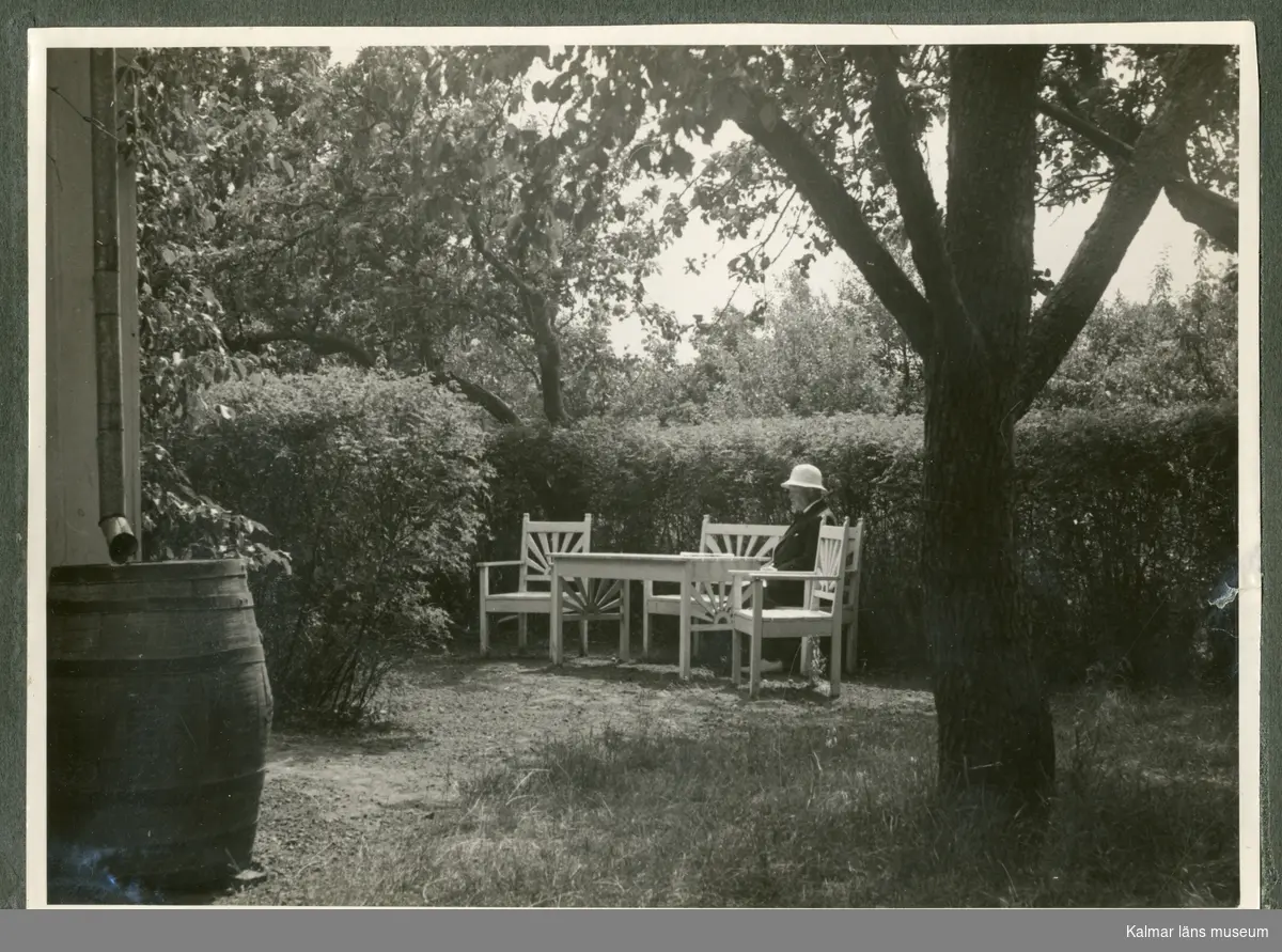 Svart-vitt fotografi som föreställer en man klädd i vit hatt som sitter i vita trädgårdsmöbler i en trädgård. Troligtvis Waldemar Swahns sommarbostad på Stångudden, Ekenäs.