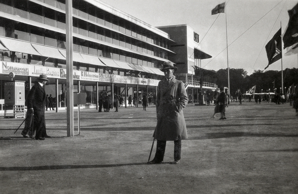 En man i rock och hatt står framför en långsträckt utställningsbyggnad på Stockholmsutställningen 1930.