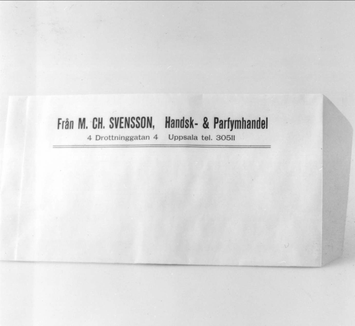 Påse av beige-rosa papper med tryckt blå text "Från M Ch. Svensson, Handsk & Parfymhandel, 4 Drottninggatan 4, Uppsala, tel. 305 11".

