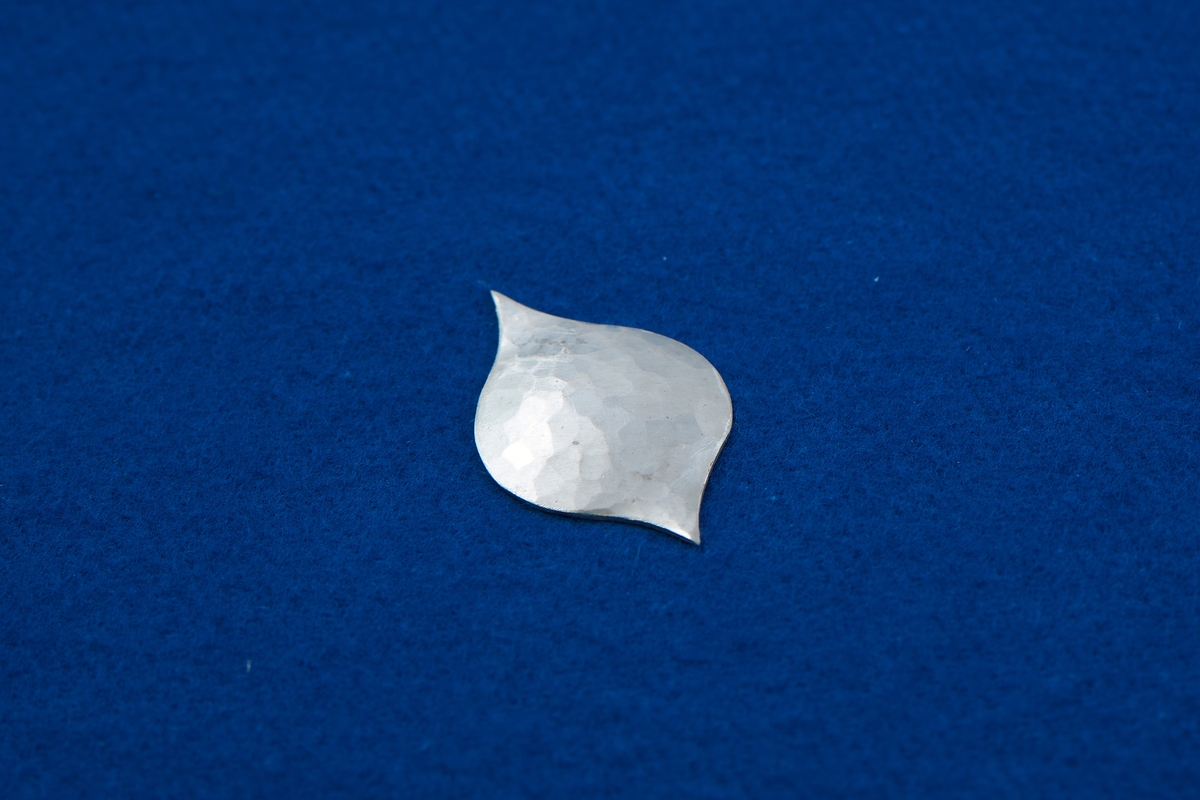Silverplåt i droppform med två spetsiga ändar på vardera sida. Plåten har lätt konkav böjning och hamrad yta.