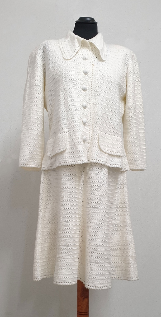 Hvit heklet drakt bestående av ermeløs kjole og jakke. Jakken har knapper i front, pyntelommer på hvert forstykke, skjortekrage og skulderputer. Flekk i front på både jakke og kjole.