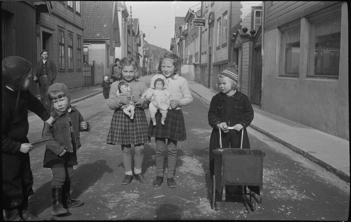 Barn i en gate i Egersund sentrum. To av jentene viser frem dukkene sine og ei har dukkevogn med. I bakgrunnen er Viktoria hotell. Bilde nr.2 er av en mann med fotoapparat