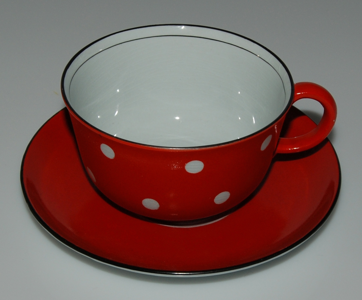 Tekopp med fat, röd glasyr med vita prickar samt två svarta linjer på insidan av koppens överkant. Okänd formgivare och dekoratör. Kan vara inspiration till den senare (1949-57) av Helmer Ringström skapade dekoren Amanita.