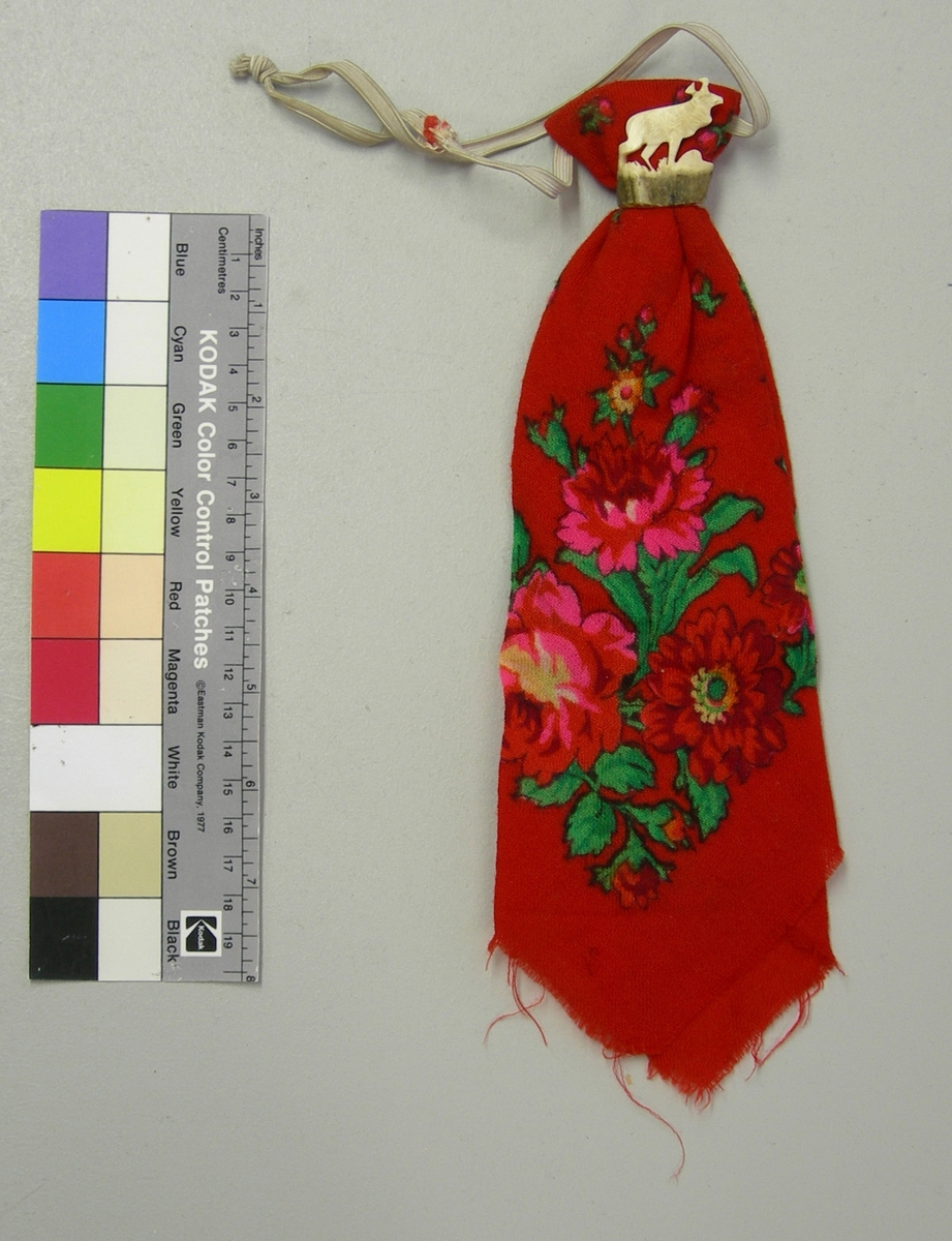 Röd tunn yllescarf i treskaftad kypert. Blomstertryck i grönt, rött, cerise, gult, orange och svart. Vikt för att bäras som slips; sölja i horn i form av en kronhjort och resårband för att fästas runt halsen. Ofållad.
