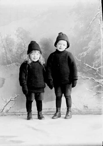 Grupporträtt med två små pojkar i stickade tröjor, mössa och vantar. De håller varandra i handen och står framför scenografi med vinterlandskap.