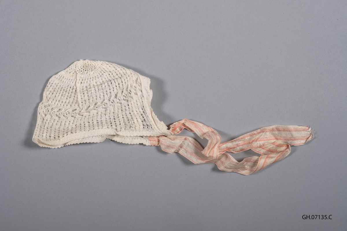 Dåpslue i hvitt bomullsgarn. Trolig strikket. Snurpet og sydd sammen på baksiden. Strikket kan sydd på i nakkelinningen. Originale knytebånd i silke, rosa og hvit-stripede.