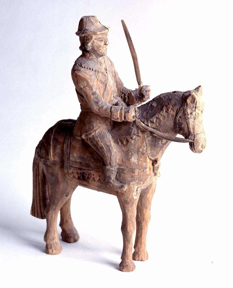 Figur av tre, forestiller hest med rytter. Mannen har hatt og sverd i hånden, og hestens seletøy i den andre hånden.