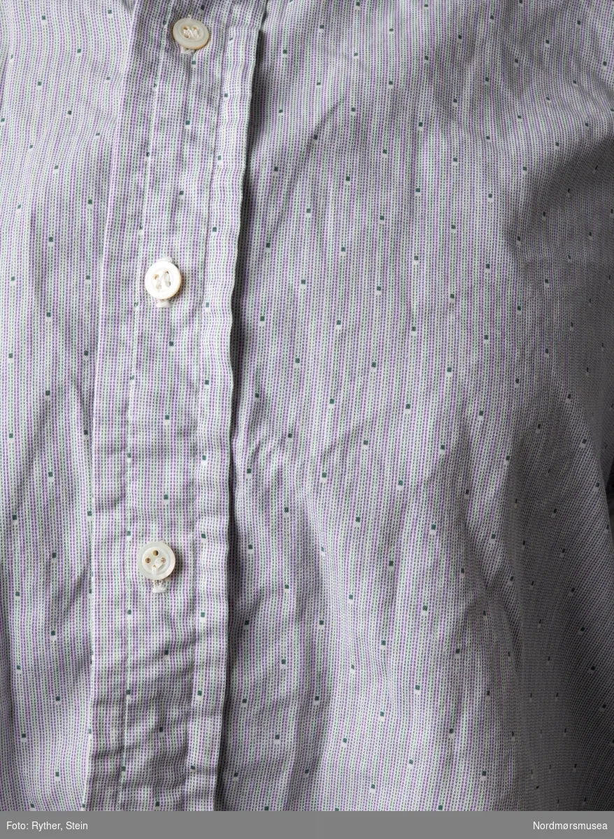 Herreskjorte med stripete mønster, rødt og blågrønt på hvit bakgrunn, med jevnlige små hvite felt med små blågrønne firkanter. Halslinning med tre knapphull for feste av løs snipp. Knappestolpe foran. Franske mansjetter og hull for mansjettknapper.