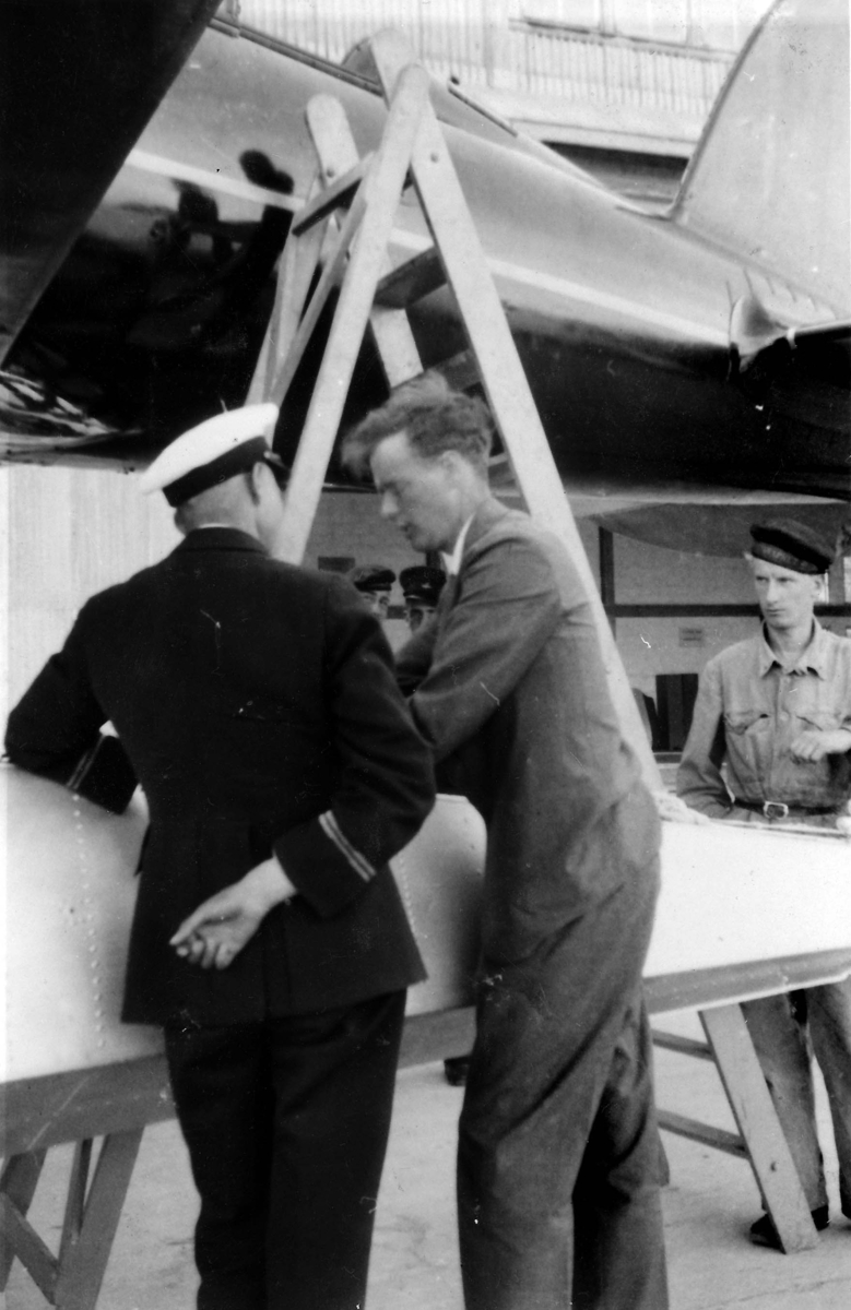 Charles Lindbergh besöker F 2 Karlskrona flygstation på Stumholmen i september 1933. Lindbergh i samtal med en svensk militär vid ett flygplan. I bakgrunden syns en man ur flottan.