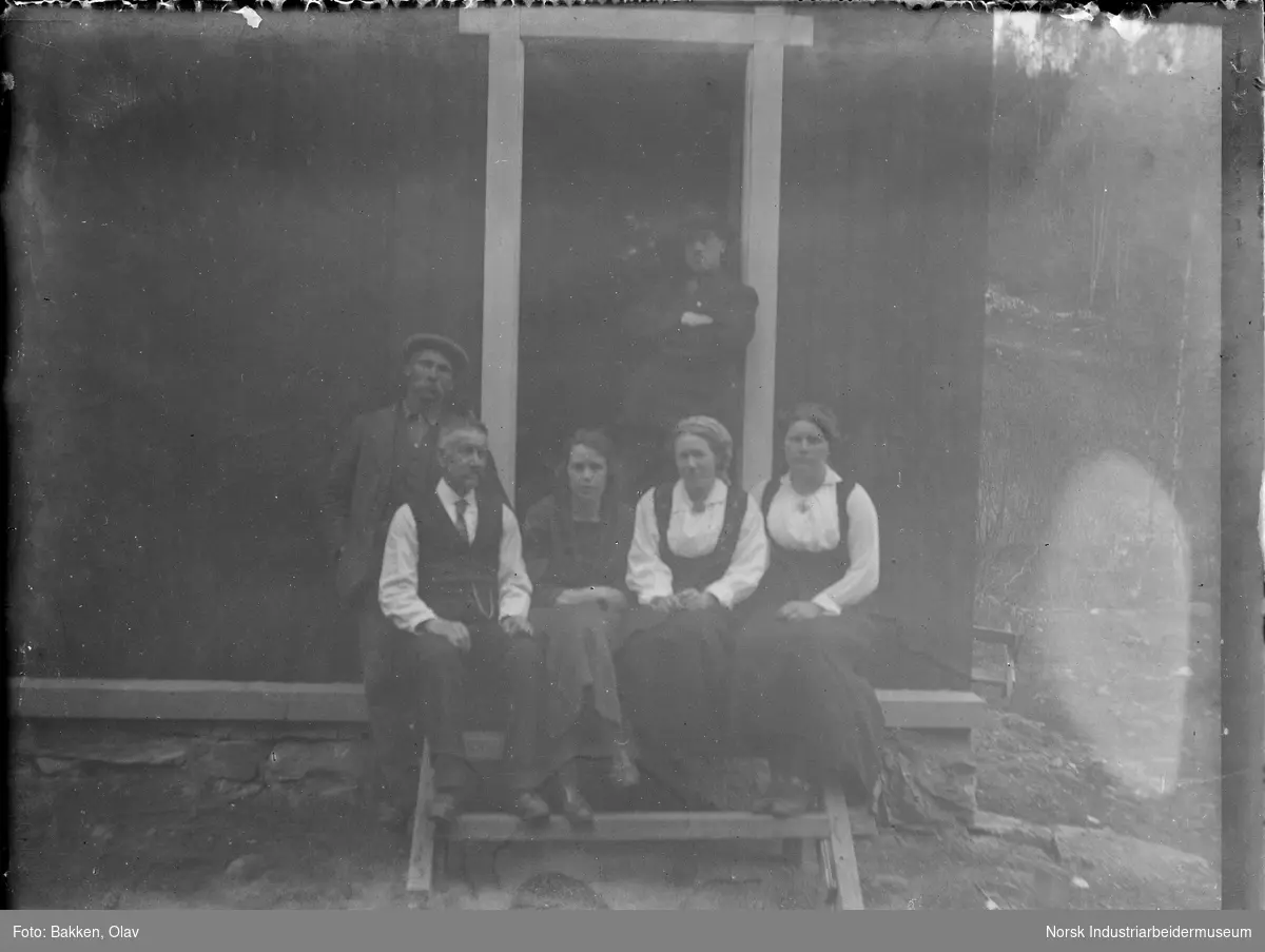 Tre menn og tre kvinner ved inngangstrappen på et hus. Kvinnene og en mann sittende på trappen, en mann stående ved siden av og en mann stående i inngangsdøren.