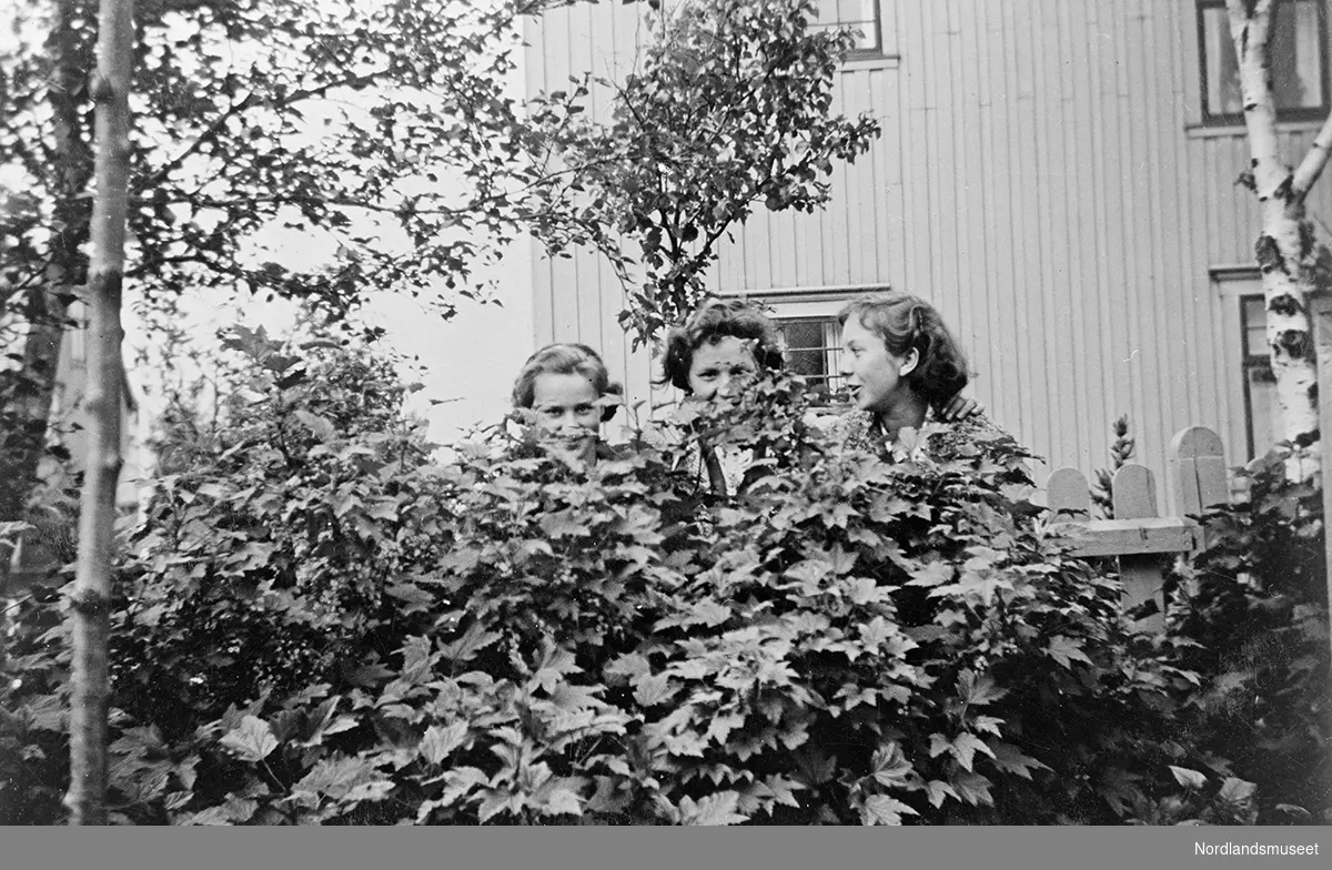 Anna Kristine Vatten, datter til Helga og Ole Vatten, sammen med 2 andre venninner. Personene er delvis gjemt bak noen planter. Litt av et bolighus vises i bakgrunnen.