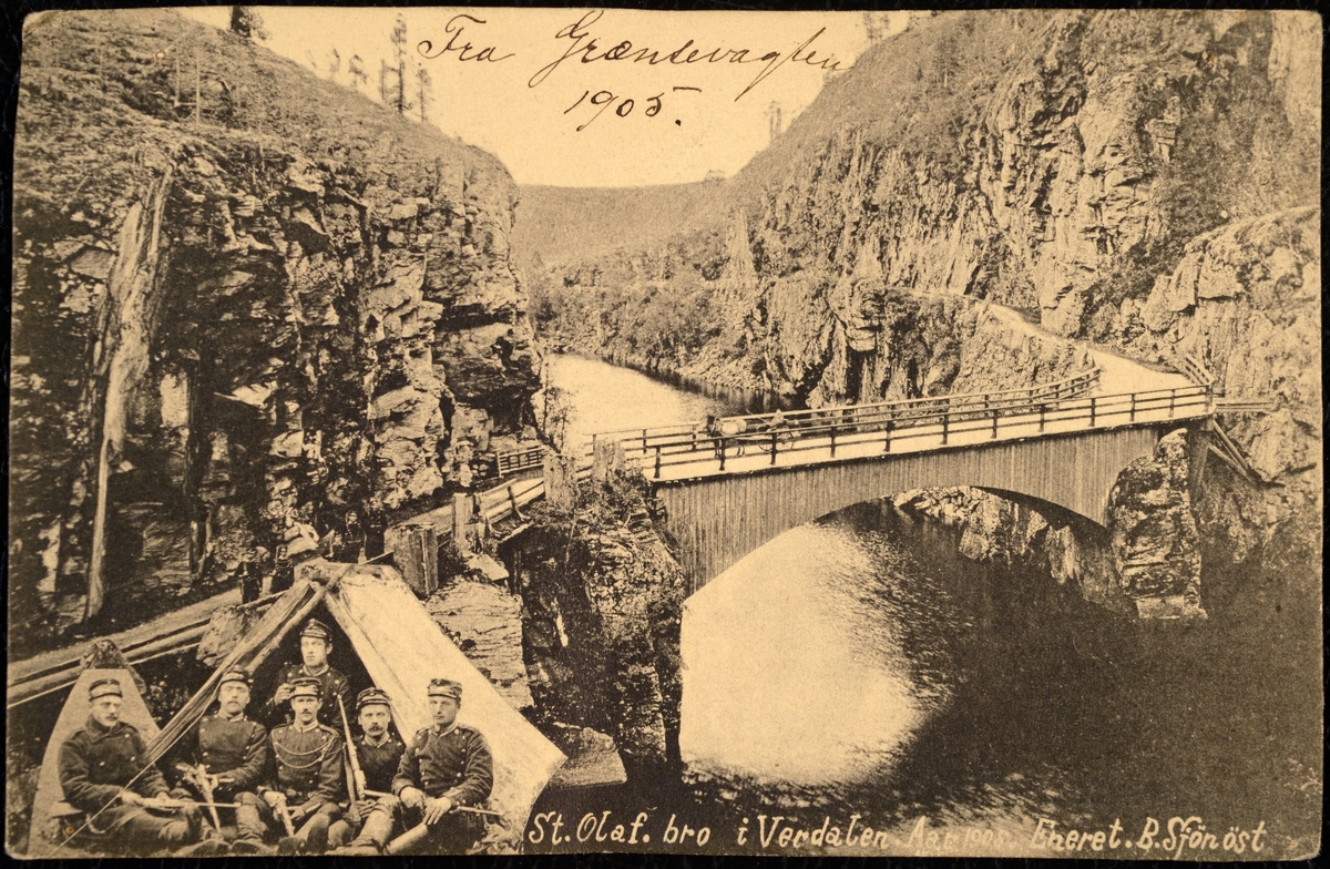 Postkort med bilde fra St. Olaf bro i Verdalen. Sort hvitt bilde av broen og 6 soldater nede i venstre hjørne på bildet. De sitter utenfor et telt. Håndskrevet tekst øvert på kortet. Og orginaltekst nederst på kortet. Trykt tekst på baksiden.