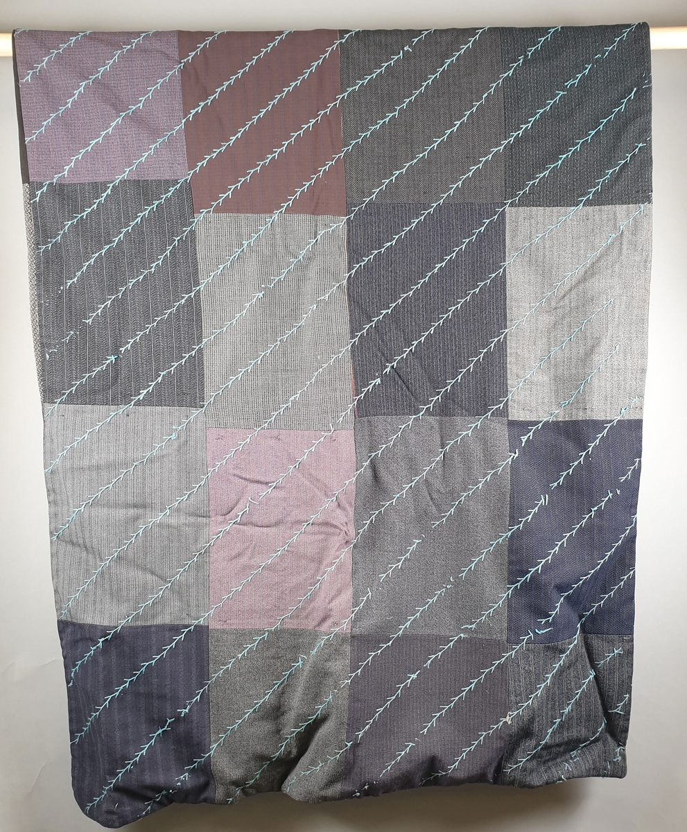 Lappeteppe satt sammen av flere stoffstykker av ull. Teppet er dobbelt og på forsiden er det brodert diagonale striper med lyseblått ullgarn.
