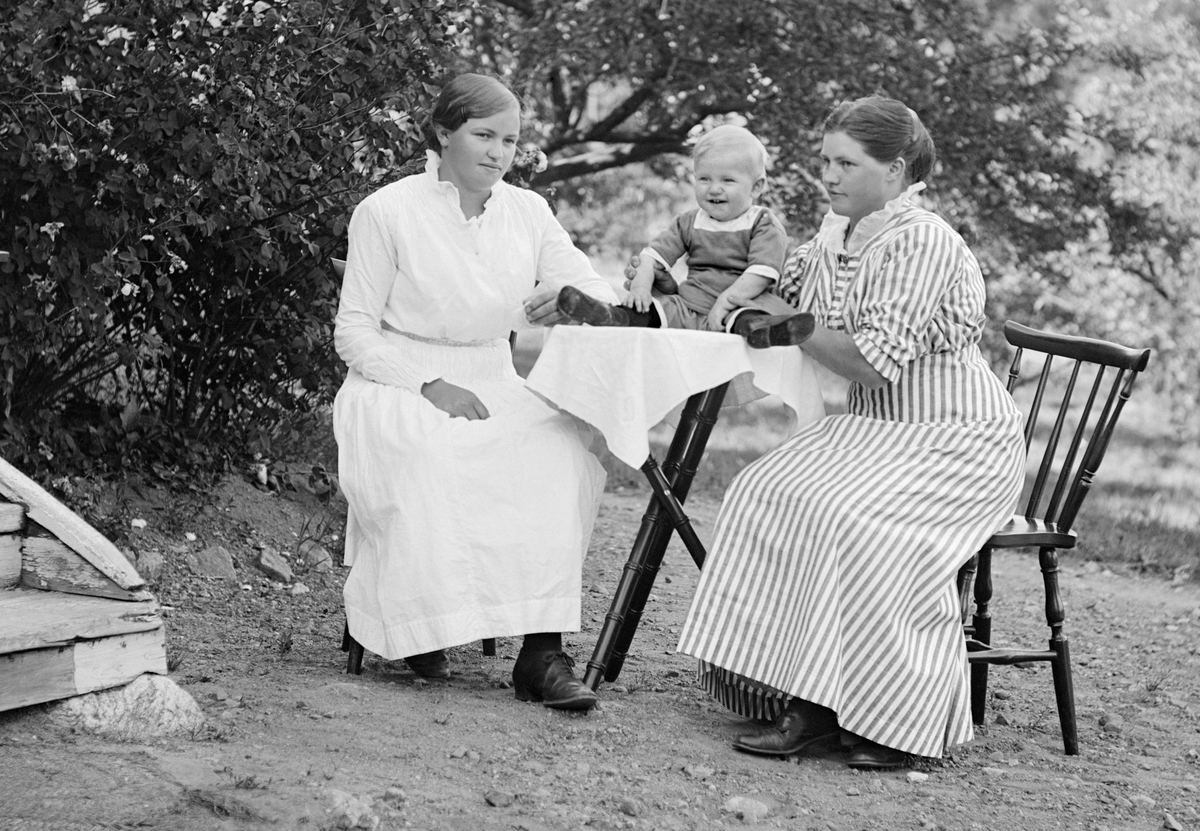 Två väninnorna, varav Magda Axelsson till höger är identifierad, låter sig förevigas i fotograf Emil Durlings improviserade friluftsateljé i sin trädgård till hemmet Strömmen i Sankt Anna. Tiden är sommaren 1917 så rimligtvis är barnet Magdas förstfödde, årsgamla son.