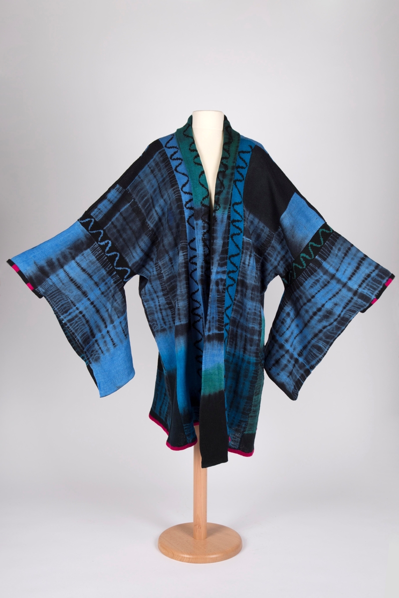 Lang, strikket damejakke i primærsnitt, kimonoinspirert i sin konstruksjon. Innfargingen av garnet, og til dels mønsterstrikkingen, gir jakken et preg av ikatvev. Bolen er sydd sammen av lange remser, og skulderbredden så bred at den henger langt ned over overarmene. Ermene fremkommer ved enda en remse, sydd på bolen nedenfor skuldrene, og atskilt fra denne ved smale bånd med innstrikket bølgemønster. Fra ermene henger lange flak, som på en kimono, men selve åpningen ved håndleddene er mye smalere, og har en påsydd, smal kant med svarte og røde striper. Også jakken har en smal kant nederst. Denne er hovedsakelig rød. Jakkens front er åpen og rundt hals og ned langs åpningen er påsydd en ny remse, delsvis nedbrettet som krage og slag. Også den med bølgeborder i mønsterstrikk. Denne er ekstra lang og henger som smale flak nedenfor jakkens øvrige avslutning. Ryggstykket er sammenføyd av de samme remsene, og noe lengre, ned i spiss, midt bak. Bolens og ermenes mønster preges ellers av store rutefelt fremkommet ved batikkfarging. Jakken er hovedsakelig blå og svart, men stedvis med overganger til mer turkise og grønne partier i krageområdet.

Luen har dobbel oppbrett, ensfarget blå. Pullen er rund, med bølgebord i mønsterstrikk.