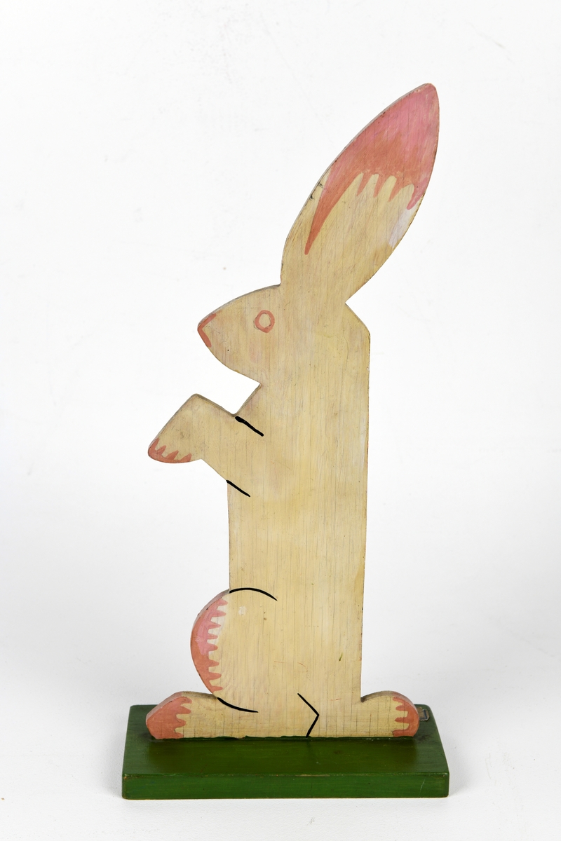En harefigur laget av tynn treplate. Haren sitter oppreist med forlabbene foran seg. Figuren er malt lys rosa med mørkere rosa på ører, labber og mage. Snute og øye er markert med mørk rosa. Den er montert  på en grønnmalt treplate.