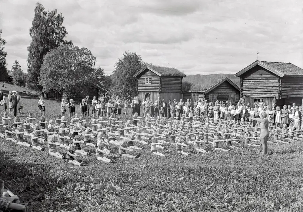 Barn sitter i rekker og øver på svømmetak etter instruksjon fra svømmelæreren. Rundt står foreldrene og ser på. Museumsbygninger i bakgrunnen.