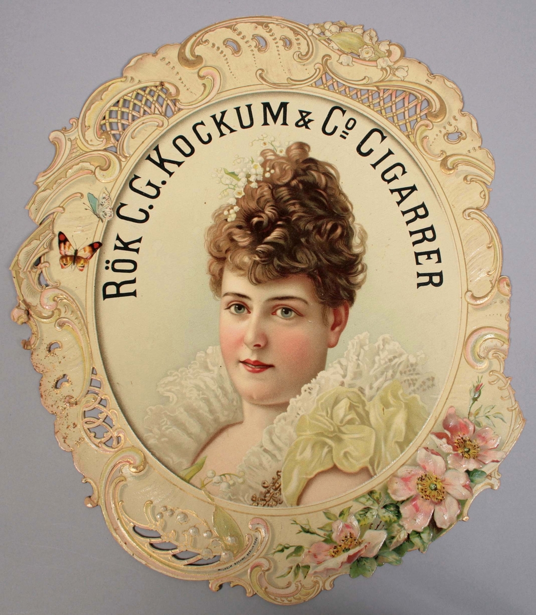 Pressad papperskylt med tryckt dekor. I form av "ansiktsbild av kvinna med uppsatt brunt hår och vita klockformade blommor. Klädd i armlös vit spetsklänning, med höga rynkor och pastellgröna rosetter på vänster axel". Ovanför hennes huvud texten: RÖK C.G. KOCKUMS & Co CIGARRER. 
Allt inramat av en oval ram i nyrokoko, med inslag av blommor och fjärilar i pastellfärger. Märkt: Wilhelm Boehme, Berlin