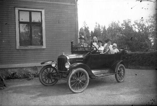 En öppen bil står parkerad vid en husknut. En man med hatt står på andra sidan bilen. I bilden sitter en kvinna i baksätet med tre barn. En något äldre pojke med keps sitter vid ratten.