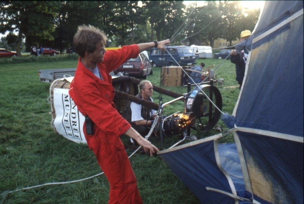 Två personer fyller en luftballong med varmluft från en brännare. En sitter på marken och en håller i ballonghöljet. På ballongkorgen står det "Andréemuseet".
