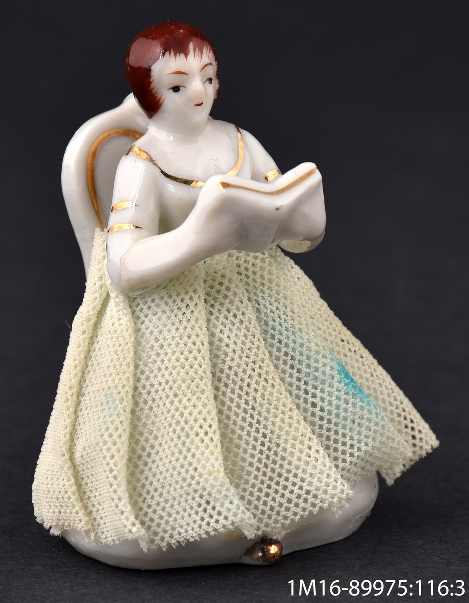 Leksak av porslin, i form av en sångerska som sitter på en stol i vitt och guld. Sångerskan håller en bok i sina händer. Kjolen är gjord av veckat tyg.