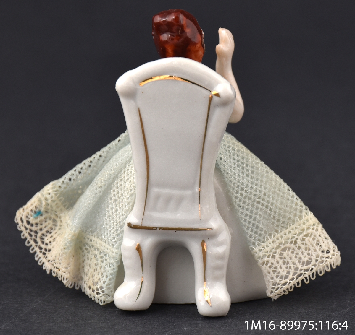 Leksak av porslin, i form av en dam som sitter på en stol i vitt och guld och applåderar. Damens kjol är gjord av veckat tyg med en spetskant nedtill.