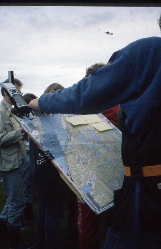 En man fotograferad snett bakifrån. Han håller i en karta fastsatt på en tunn träskiva. På kartan sitter gula post-it lappar och en kommunikationsradio fastspänd i över vänstra hörnet.