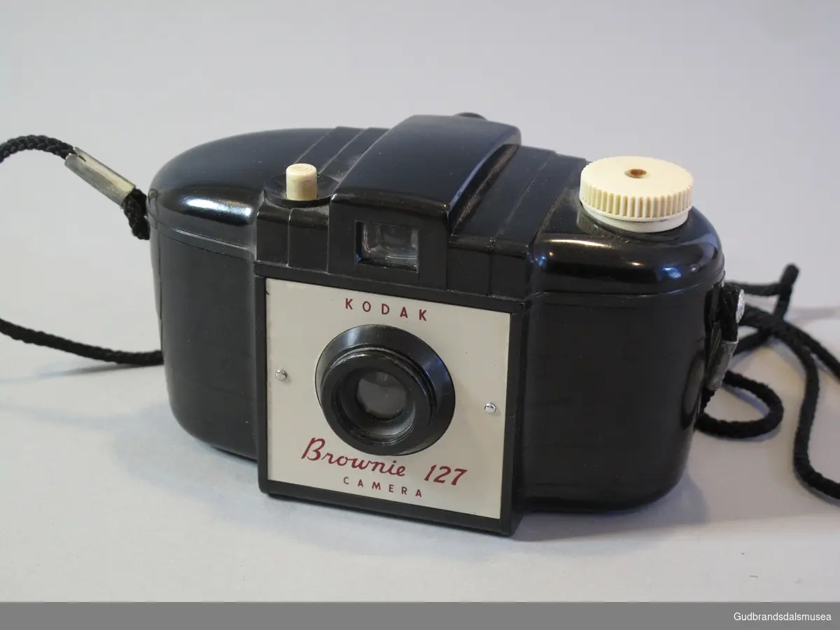 Kodak Brownie 127. Sort plastkamera for 127 mm film. Oval form. snor til å feste kameraet rundt halsen. Kameravekse i brunt lær med reim og metalllås.