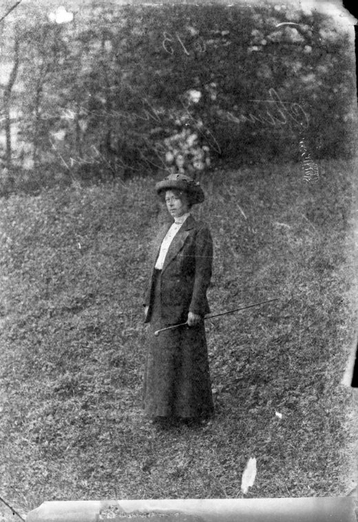 Porträtt av en kvinna som står på en gräsmatta. Hon står lite i profil och håller en käpp i vänster hand.