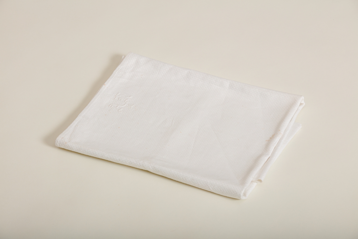 Håndkle av hvit lin med mønstereffekt i vevningen. Maskinsydd fold i hver ende. Hvitt håndbrodert monogram i plattsøm. Påsydd hank av bomullsbånd.