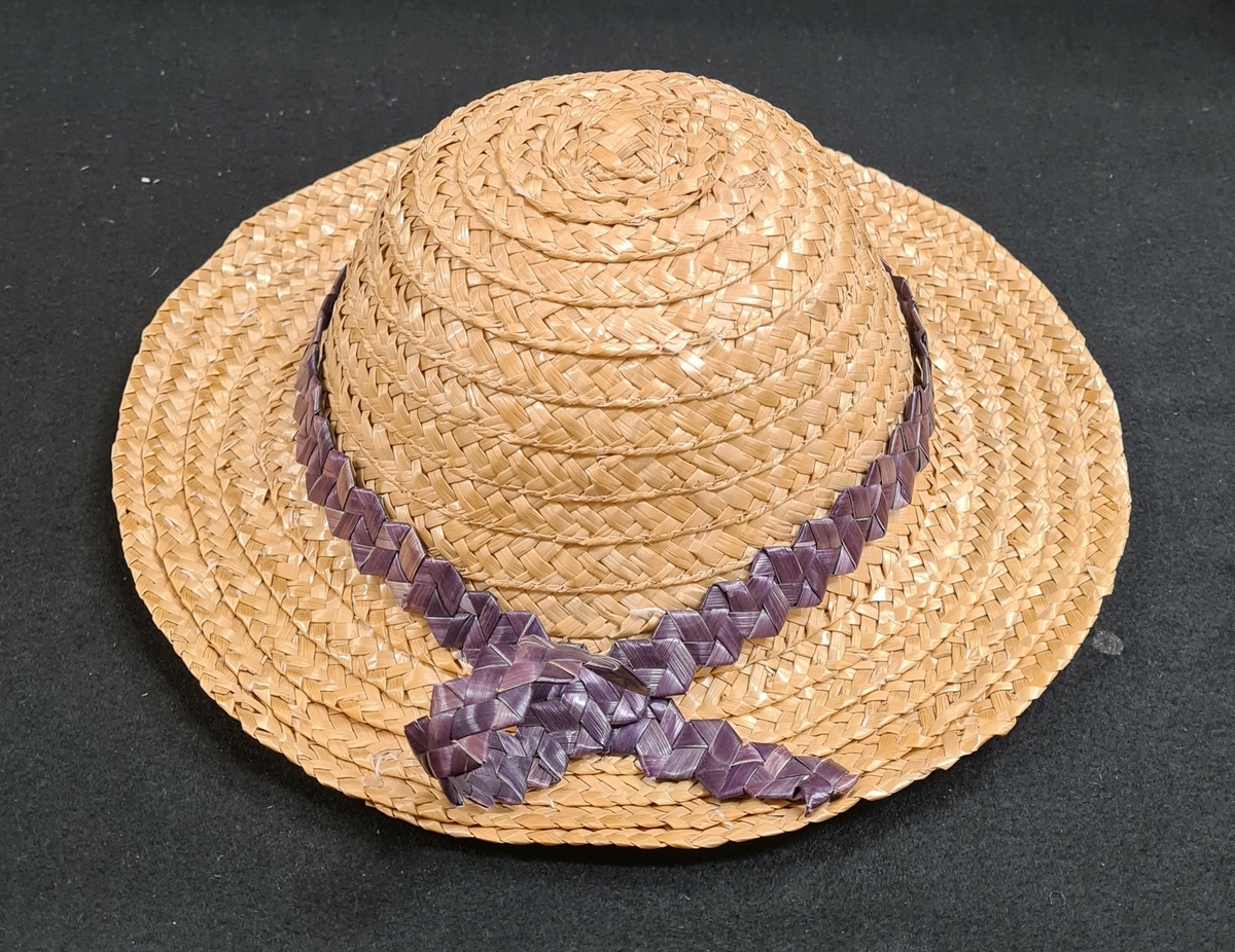 Halmhatt med kulle och brätte. Hatten är försedd med ett lila, taggflätat band. Hatten är maskinsydd.