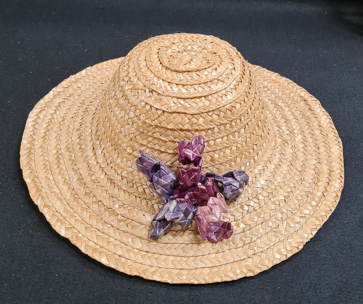 Halmhatt med kulle och brätte. Hatten är försedd med ett lila, taggflätat band formad som blomma. Hatten är maskinsydd.