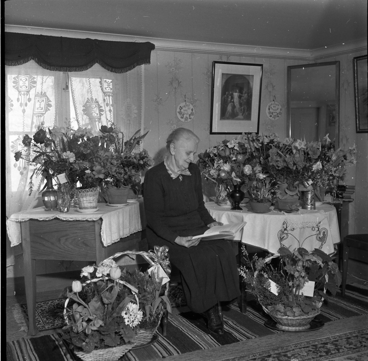En kvinna sitter omgiven av blombuketter och blomsterarrangemang. Hon håller i en bok. Hon bör en svart klänning med spetskrage.