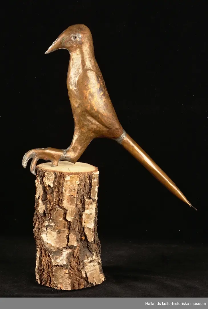 Skulptur av koppar föreställande en fågel. Skulpturen står på en plattform av björk.