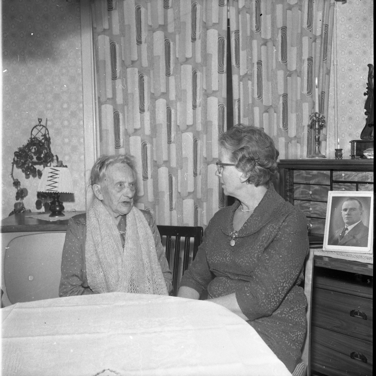En äldre dam sitter i en hemmiljö och pratar med en något yngre dam. Ett porträtt av en man står på chiffonjen bredvid dem.