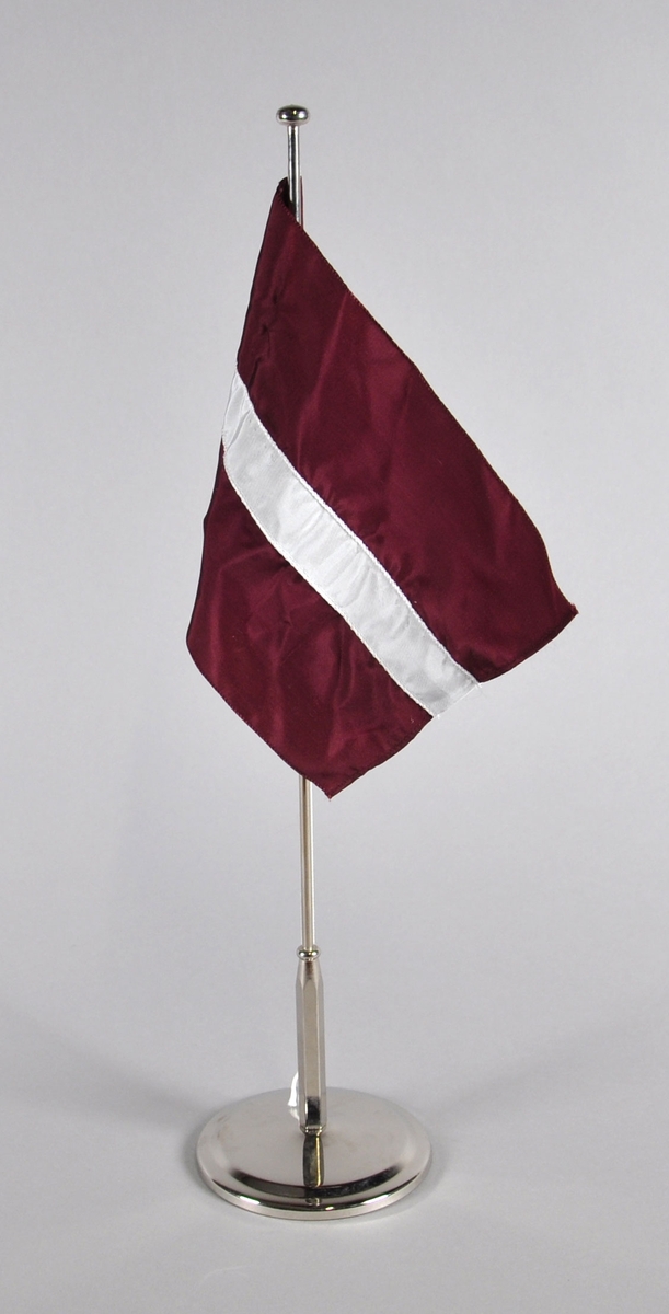 Bordflagg på stang fra Latvia. Tre horisontale striper, fiolett øverst og nederst og en smal hvit stripe på midten.