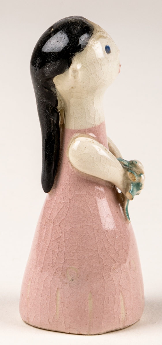 Brudtärna till figuringrupp Brudfölje. Tärnan har mörkt hår och rosa klänning. Formgiven av Dorothy Clough 1955, Gefle Porslinsfabrik. På undersidan svaga rester av stämpeltryckt logga med GEFLE och tre rundugnar.