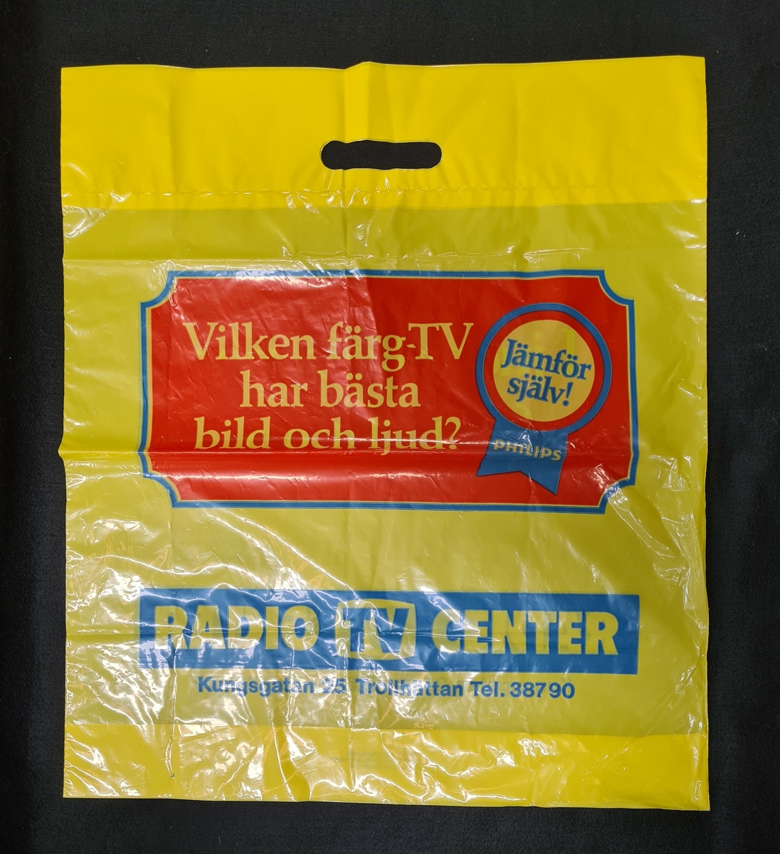 Plastkasse, gul, med logga från Radio TV center i Trollhättan. 

På kassen står:
Vilken färg-TV har bäst bild och ljus?
Jämför själv!
Philips
 
Radio TV center
Kungsgatan 25 Trollhättan Tel. 38790