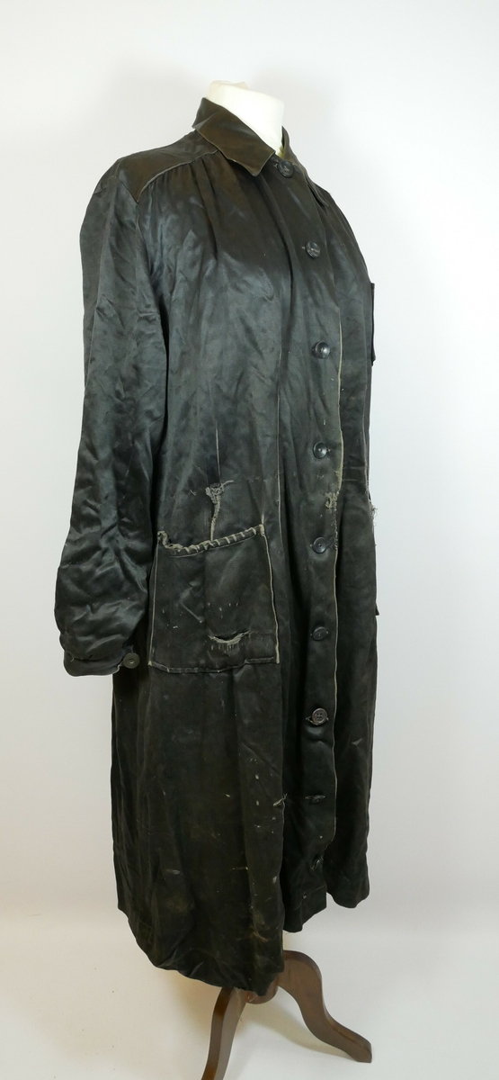 Knelang svart jakke i silkelignende tekstil. Jakken har en krage og ni svarte knapper foran på jakken. Den har to lommer på hver side og en brystlomme på venstre side. Hvert erme har en svart knapp