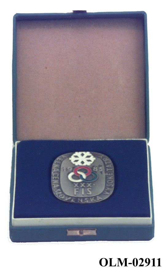 Oval minnemedalje med snøkrystall øverst, årstall under, tre ringer i rødt, hvitt og blått. Tekst rundt kanten. Medaljen ligger i et blått etui.